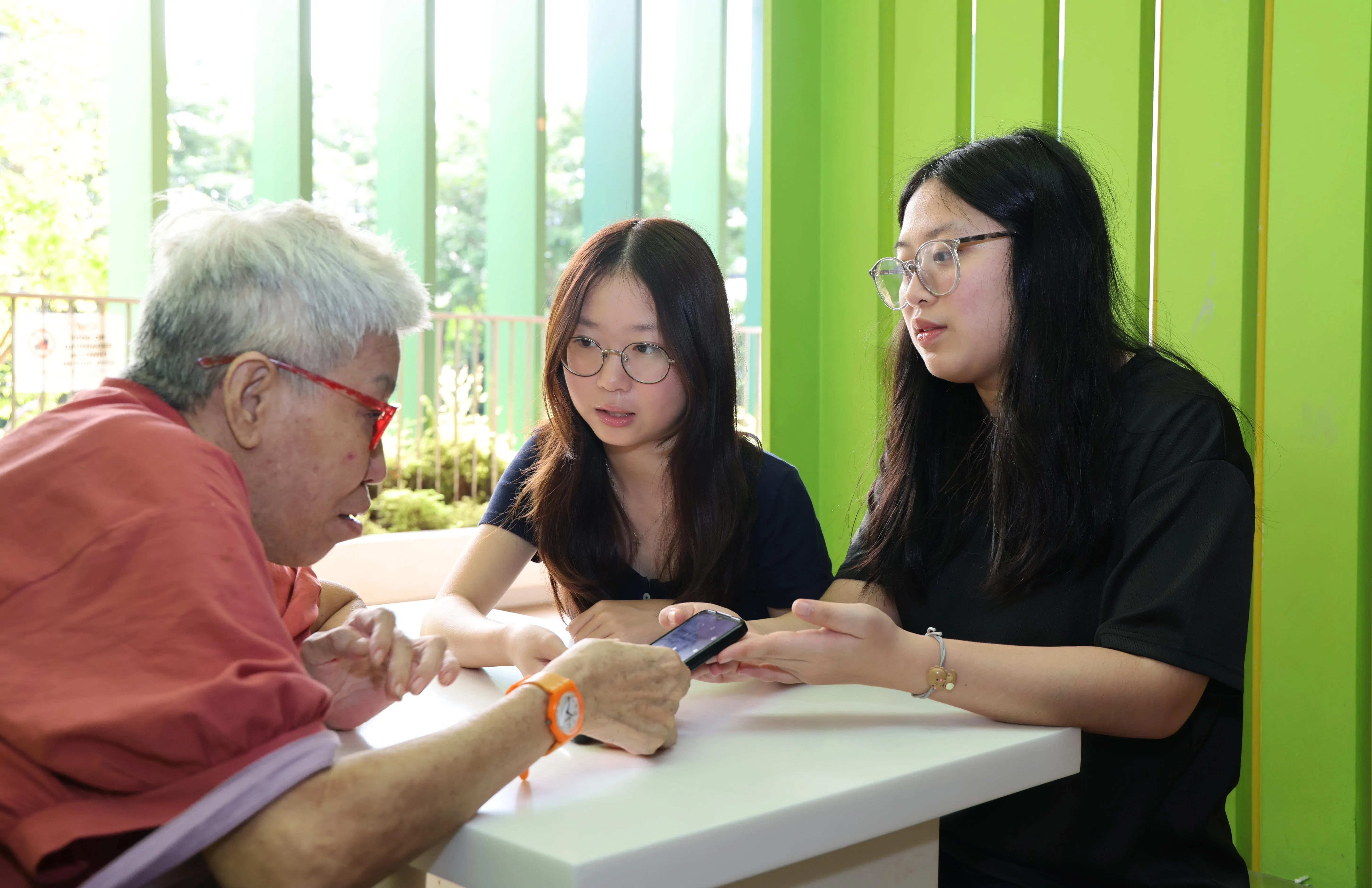 来自义安理工学院的学生义工王静宁（右起）和郑淼匀，指导义顺居民贺美莲（左）使用手机，领取社区发展理事会邻里购物券。（梁麒麟摄）