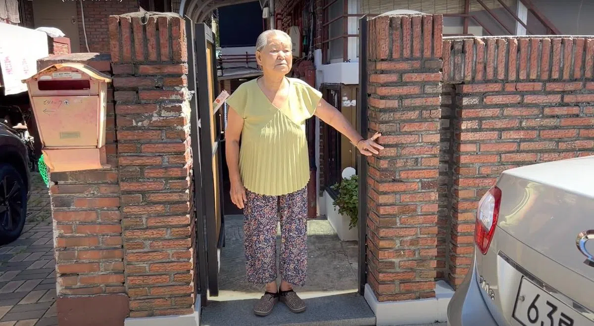 站在半地下房所在建筑门口的安淑子奶奶，她居住的房间就在身后的台阶下。由于腿动完手术后行动不便，她除了午餐到附近的老人福利馆吃，早晚餐都在家自理。她希望半地下房租约明年1月到期后，能搬进政府提供的公租房。（视频截图）