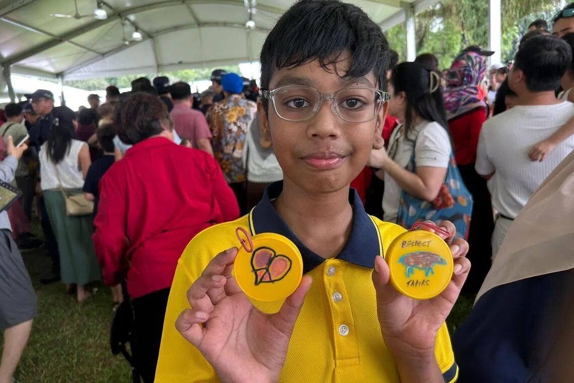 在4月19日总统府开放日上，培景小学小五学生维沙坎向记者展示总统夫妇在旧饮料瓶盖上的作画。他左手拿着尚达曼画的貘，右手拿着珍一藤木画的创意爱心。（张俊摄）