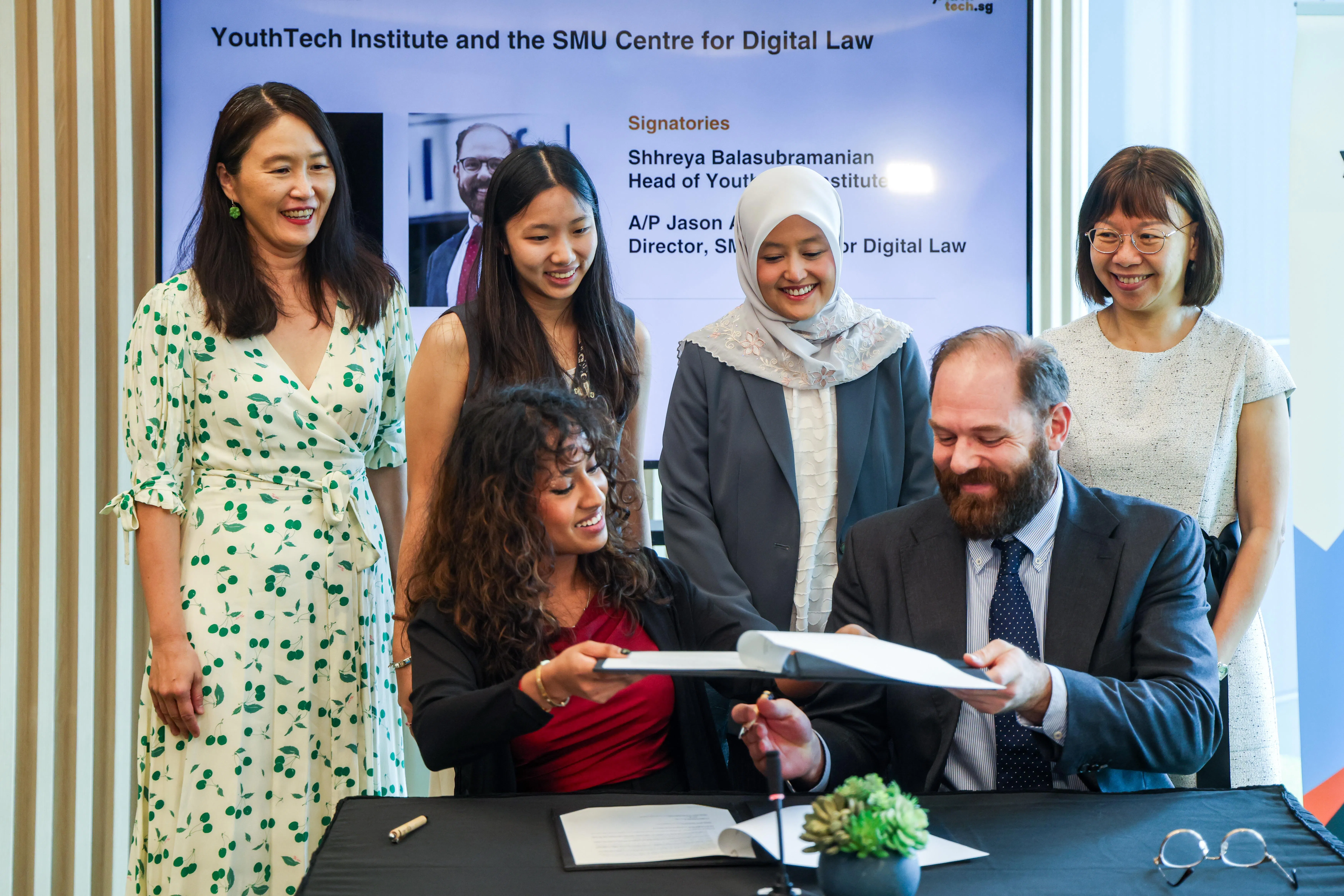 青年科技学院（YouthTech Institute）负责人什雷娅（Shhreya Balasubramanian，前排左）和新加坡管理大学数码法律中心主任兼杨邦孝法学院副教授杰森（Jason Grant Allen，前排右）签署谅解备忘录，加强青年实践与学术研究之间的交流与合作。数码发展及新闻部兼保健卫生部政务部长拉哈尤（后排右二）、新大合作与交流总监兼传播与科技学教授林珊珊（后排左起）、YouthTechSG首席影响力官兼文化与转型主管陈灵恩，以及新大杨邦孝法学院院长李佩婉教授（后排右一）见证签署仪式。（陈斌勤摄）