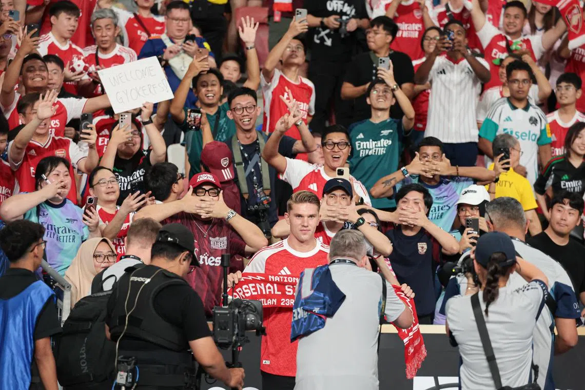 以本地阿申纳球迷为背景，吉欧克勒斯拿着球队围巾在场边拍照。后方的许多球迷，都模仿吉欧克勒斯进球后的标志性“面具”庆祝动作。（李冠卫摄）