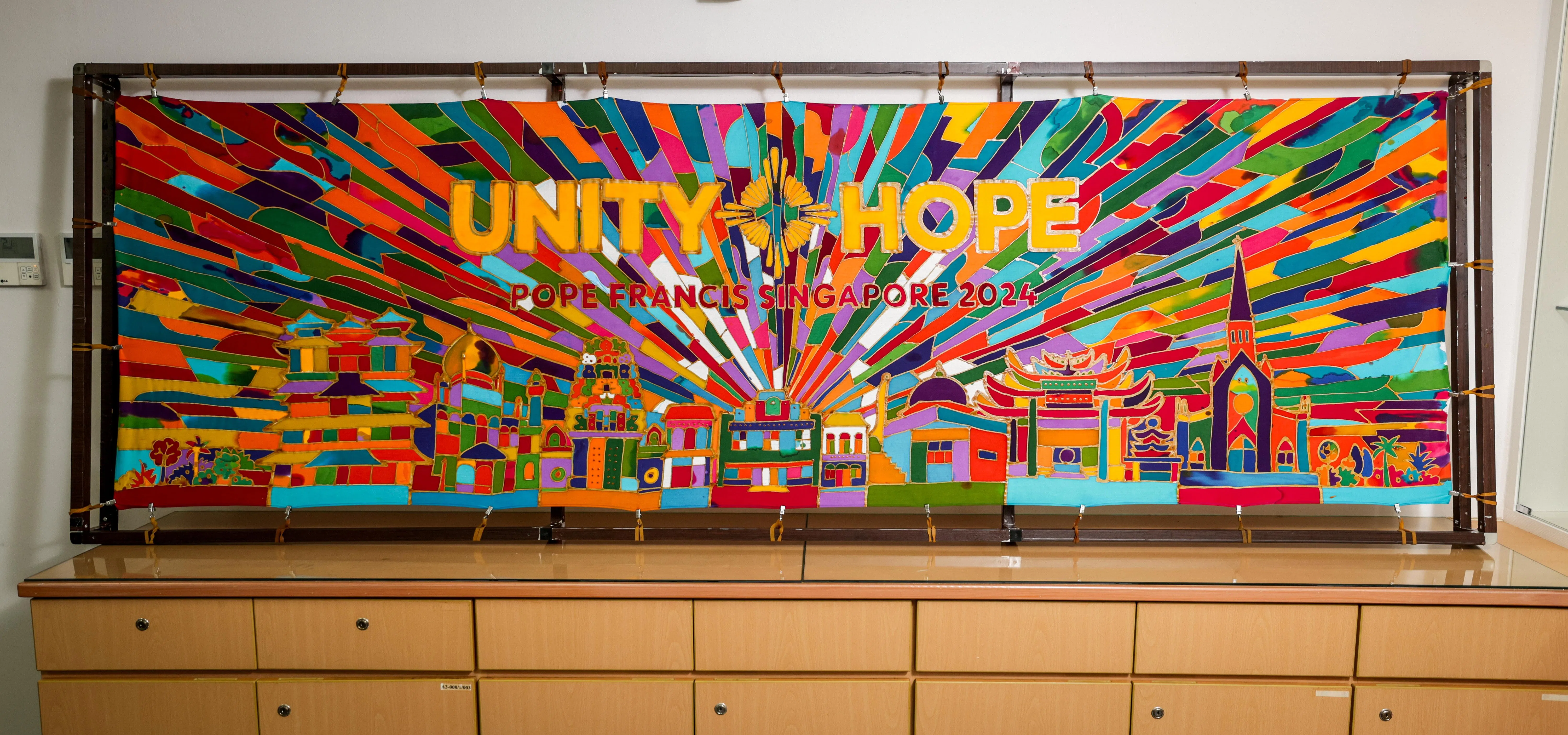 公教初院上至校长下至校工及师生共1600人，分批在一幅已勾勒出图样花纹的2.5米长布上填色。作品上方显示教宗此行主题“合一与希望”（Unity and Hope）的字样。设计也融入本地宗教场所的建筑设计，包括印度教庙、回教堂和佛寺等。（叶振忠摄）