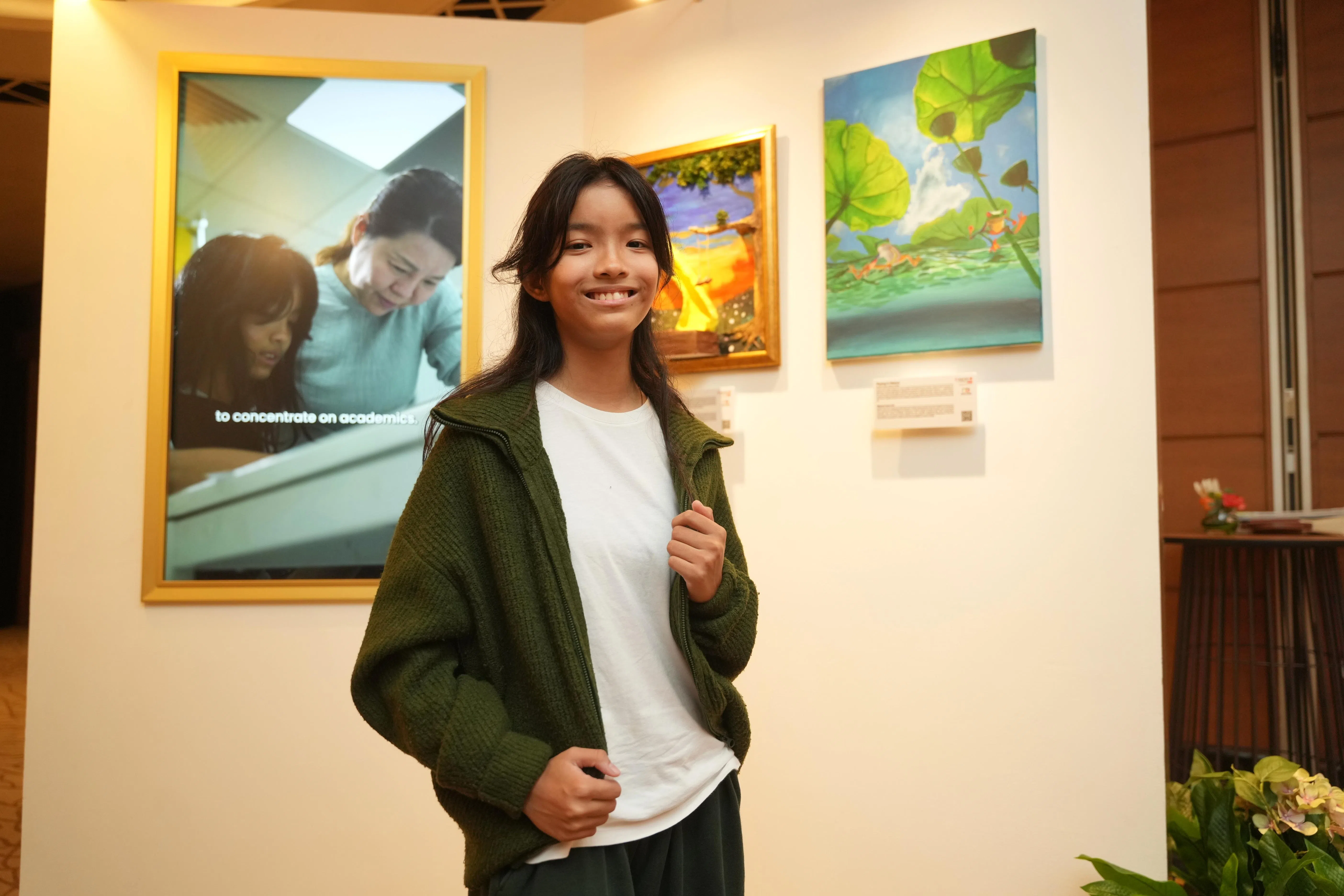 林芸仪获得“闪耀同心奖”，她在颁奖礼的展场在自己的两件作品前拍照。一件是丙烯画，描绘青蛙在莲叶间跳跃嬉戏；另一件是用粘土等材料创作的夕阳下的游乐场，描绘了放学后和朋友嬉耍的美好时光。 （谢智扬摄）