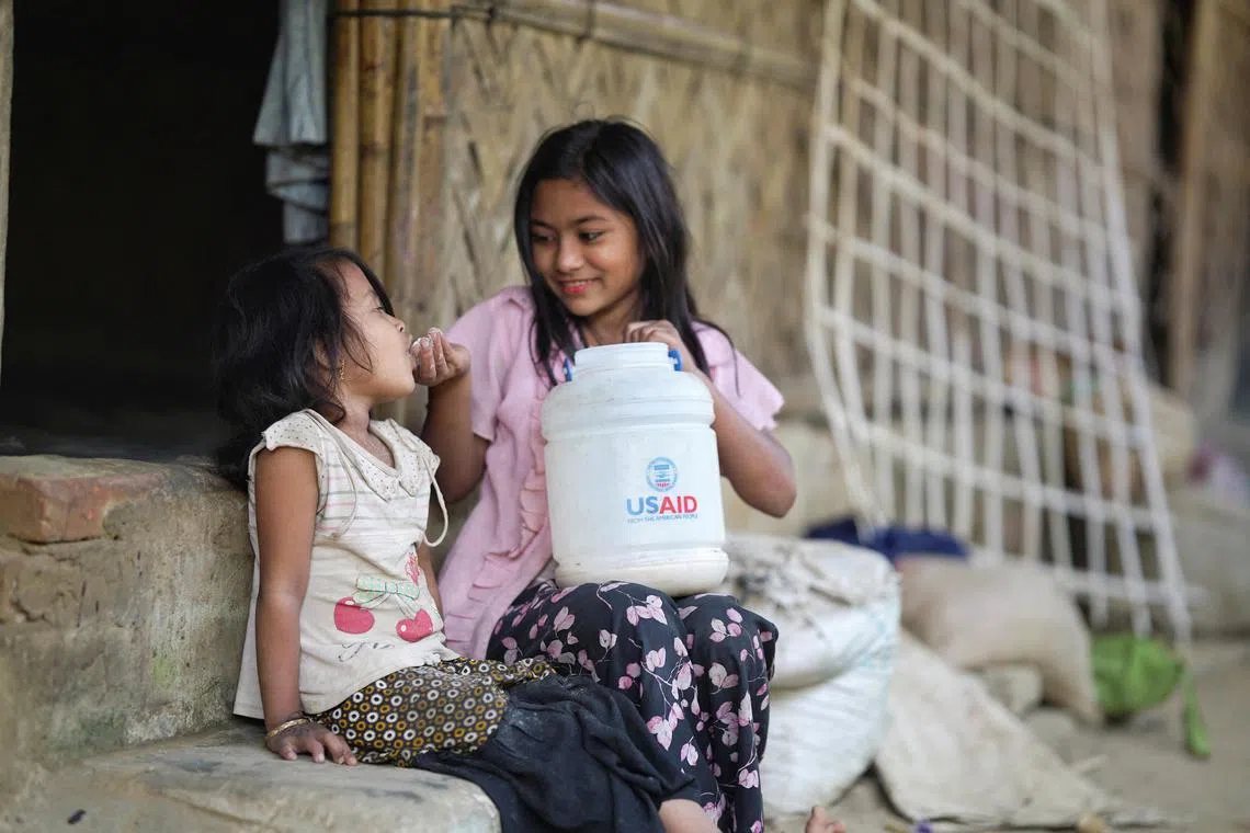 在孟加拉国的科克斯巴扎尔，一名罗兴亚女孩正在给一名幼童喂食，装食物的小桶上印有美国国际开发署名称。（路透社）