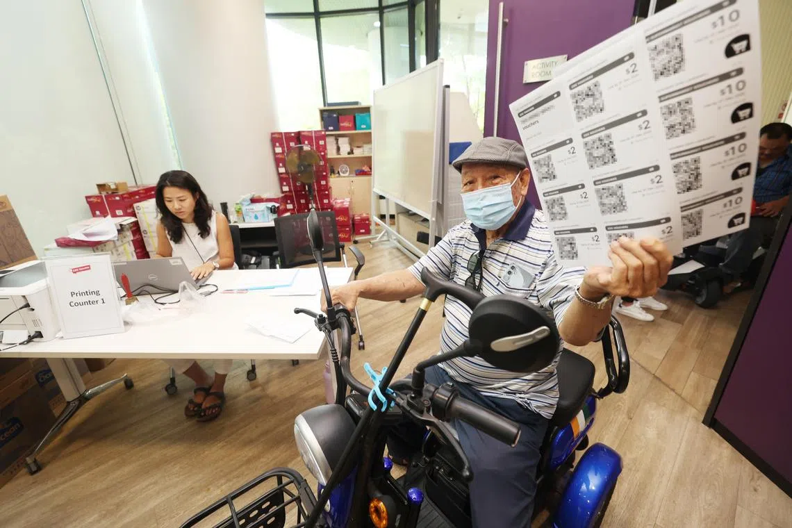 以行动辅助工具代步的退休人士张宏文（89岁，右），在义顺中民众俱乐部工作人员安排下，优先领取纸本的社区发展理事会邻里购物券，不用久等。  （郑一鸣摄）