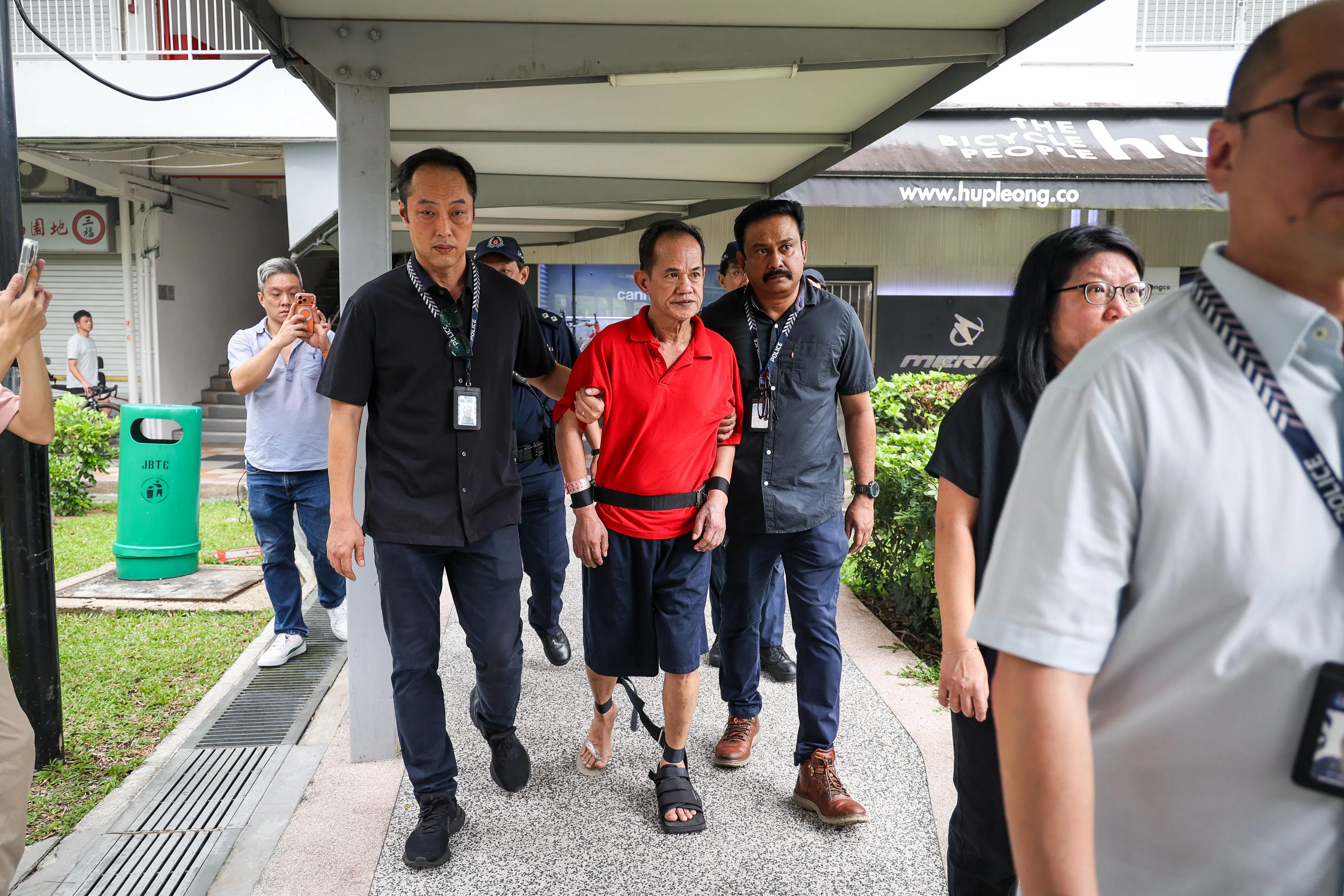 被告身穿红色Polo衫和黑色短裤，他手脚被拷，神情凝重。（张俊杰摄）