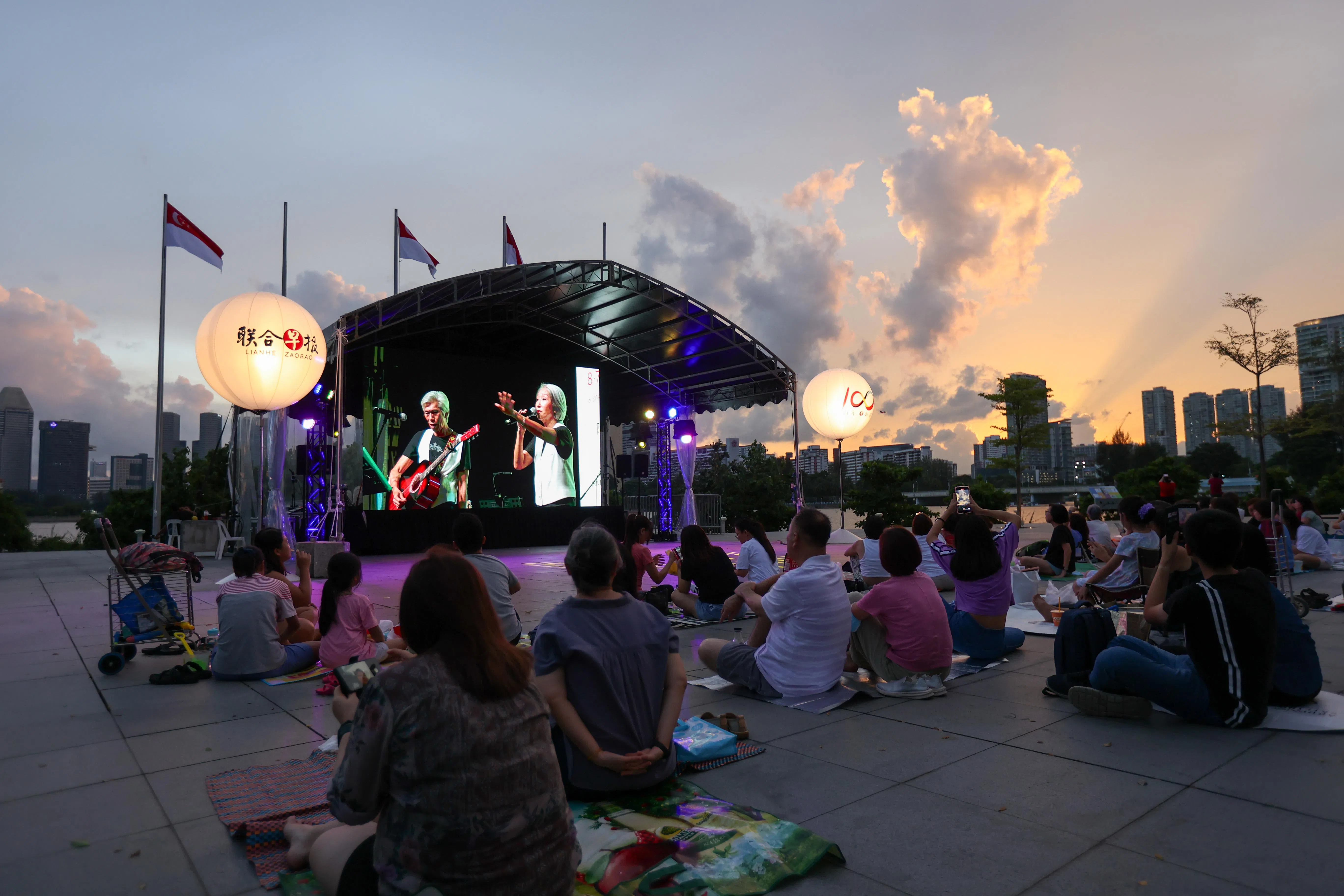 不少公众聚集在新加坡体育城的河滨走道观看现场转播。映衬着夕阳，公众随歌摆动，哼唱着熟悉的歌词。（关俊威摄）