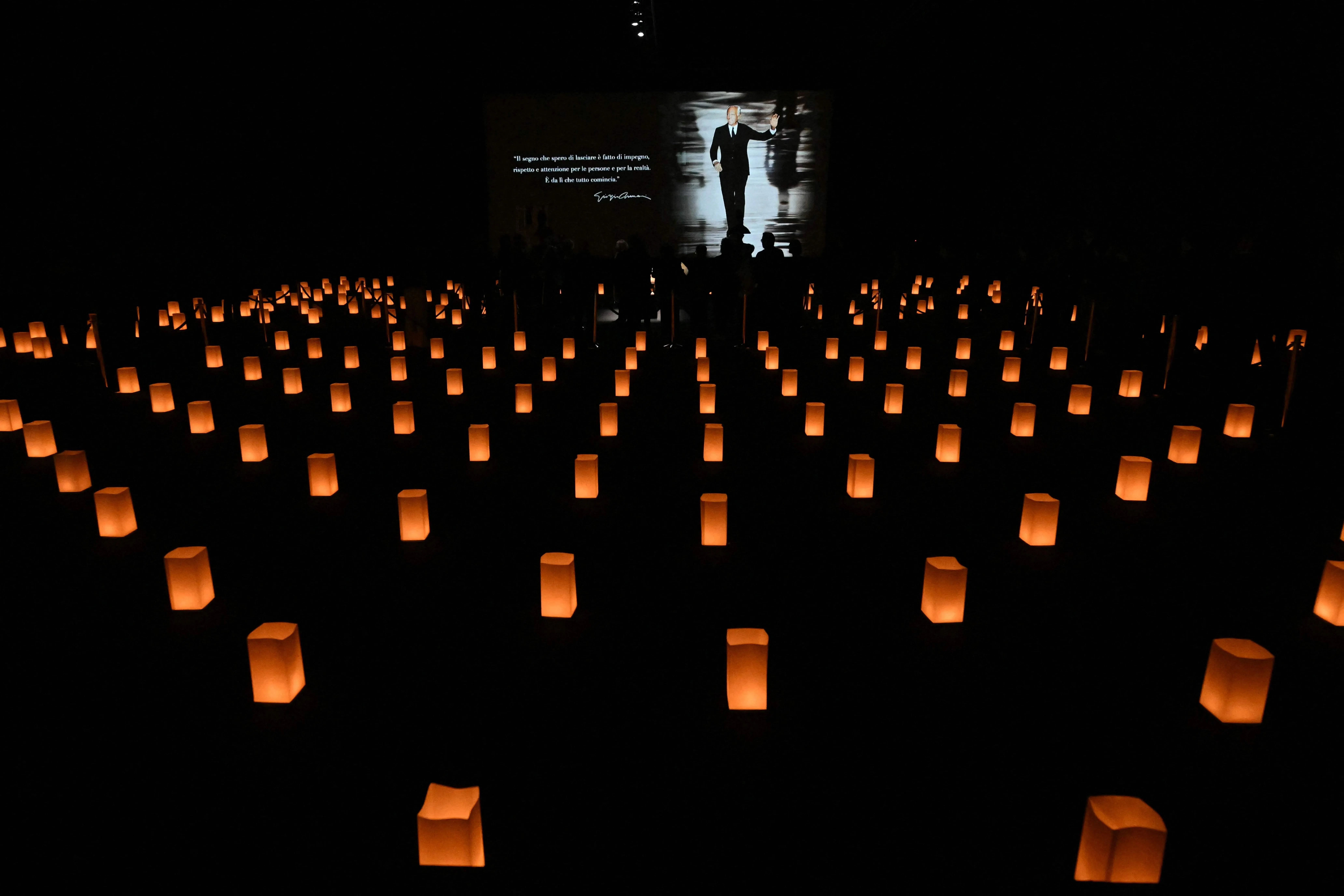 灯光昏暗的灵堂地面上排列着纸张遮挡着的蜡烛，大屏幕上播放着阿玛尼的肖像。（法新社）